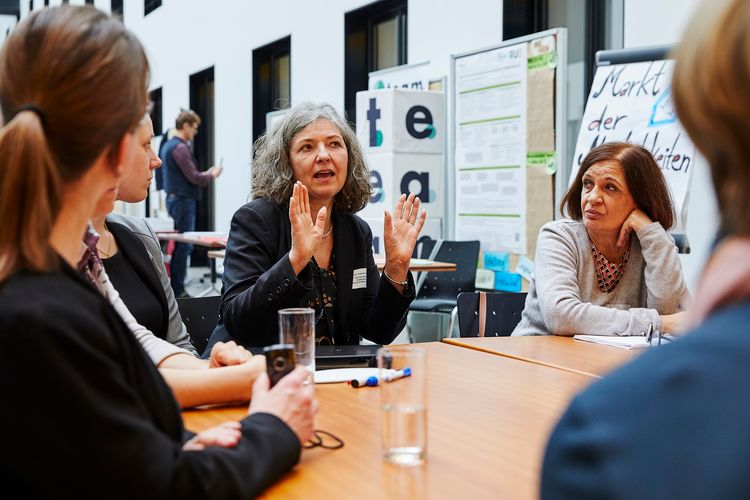 Foto: Im Vordergrund diskutieren Frauen zusammen mit der gehörlosen Professorin Fries am Thementisch. Im Hintergrund befindet sich der Markt der Möglichkeiten mit Plakaten und Sitzgelegenheiten.