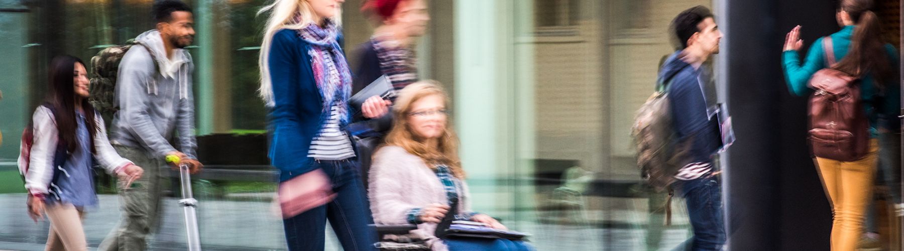 Alternativtext: Dieses Headerfoto zeigt junge Menschen mit Rollstuhl, Tretroller & zu Fuß auf ihrem Weg zu einem Hochschuleingang.